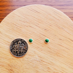 May include: A pair of small, green gemstone earrings on a wooden surface next to a US dime.