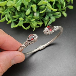 May include: A silver bangle bracelet with intricate detailing and two red gemstone accents.