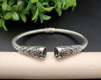 May include: A silver bangle bracelet with a twisted design and two green gemstones set into the ends. The ends of the bracelet are decorated with intricate floral patterns.