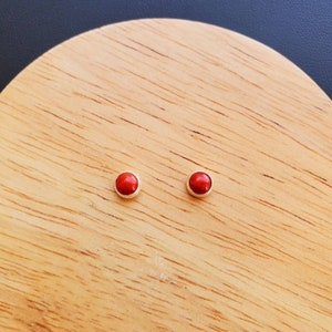 May include: A pair of small, round, red gemstone earrings with silver settings. The earrings are displayed on a wooden surface.