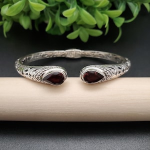 May include: A silver bangle bracelet with an ornate design and two red gemstone accents.