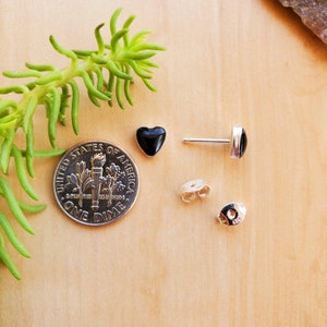 May include: A pair of black heart-shaped stud earrings with silver posts and backs. The earrings are shown next to a US dime for size comparison.