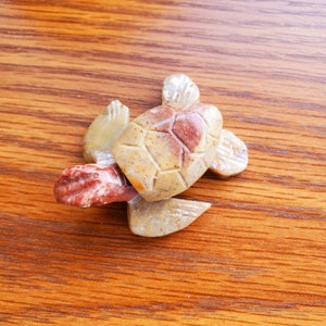 May include: A small, hand-carved stone turtle figurine. The turtle is made of a light brown stone with red and white accents. The turtle is in a swimming position with its head and legs extended.