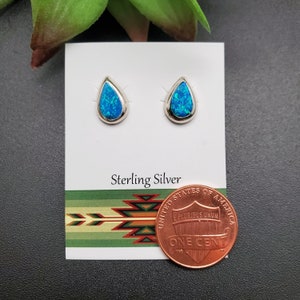 May include: A pair of sterling silver teardrop earrings with blue opal stones. The earrings are displayed on a white card with the words "Sterling Silver" printed on it. A penny is included for size reference.