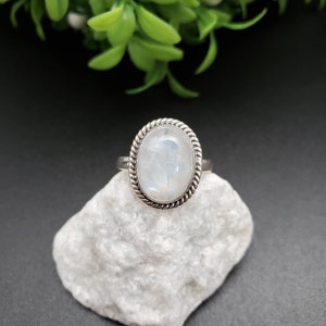 Puede incluir: Anillo de plata con una piedra lunar ovalada e iridiscente. La piedra está engastada en un bisel retorcido en forma de cuerda. El anillo se exhibe sobre una roca blanca texturizada sobre un fondo oscuro con follaje verde.