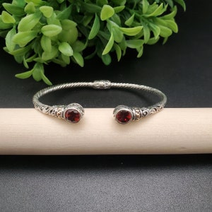May include: A silver bangle bracelet with a twisted design and two red gemstone accents. The bracelet has a delicate floral pattern.