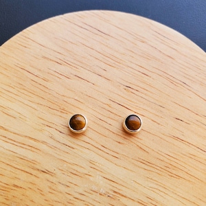 May include: A pair of silver stud earrings with round tiger's eye gemstones. The earrings are on a wooden surface.