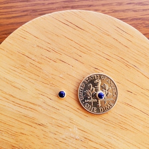 Peut inclure: Deux petites boucles d'oreilles en pierres bleues avec des montures dorées à côté d'une pièce de dix cents américaine.