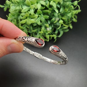May include: Silver bangle bracelet with an ornate design and two red gemstone accents.