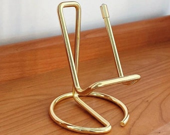 May include: A gold metal stand for a safety razor. The stand has a round base and a curved, open top.