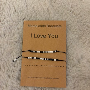 May include: Two black and white beaded bracelets with a brown string. The card reads "Morse code Bracelets I Love You" and "Morse code bracelet with a hidden message".