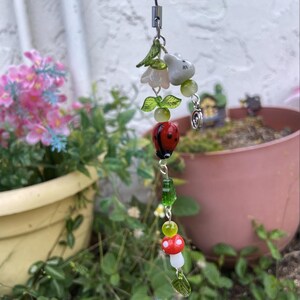 Pode incluir: Um charm caprichoso com uma joaninha vermelha, folhas verdes e um cogumelo branco com bolinhas vermelhas. O charm também inclui um coelhinho branco, flores cor-de-rosa e contas verdes, tudo amarrado em um cordão preto. Tema de jardim.