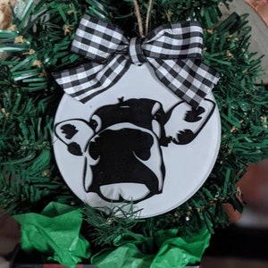 May include: A white, round Christmas ornament featuring a black and white cow face design. The ornament is topped with a black and white checkered bow and a twine hanger. The ornament is surrounded by green faux pine needles.