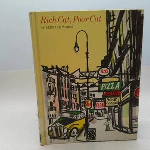 May include: A vintage hardcover book titled "Rich Cat, Poor Cat" by Bernard Waber. The cover features an illustrated city scene with a yellow car, street signs, and a pizza shop. The book's cover is yellow.