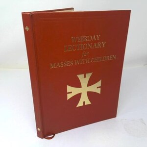 May include: A red hardcover book titled Weekday Lectionary for Masses with Children in gold lettering. The front cover features a large gold cross emblem. The spine includes a small gold cross symbol near the top and bottom edges.