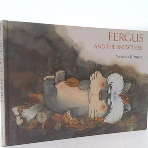 May include: A children's book cover featuring a cartoon cat with white fur on its head and a sad expression. The book is titled "Fergus and the Snow Deer" by Yasuko Kimura.