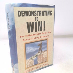 May include: A blue book with the title "Demonstrating to Win! The Indispensable Guide for Demonstrating Software" by Robert Riefstahl. The cover features an illustration of a person walking across a dollar bill bridge over a gap between two cliffs.