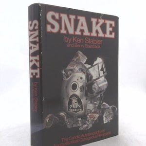 May include: A hardcover book titled "SNAKE" by Ken Stabler and Berry Stainback. The cover is black with the title in large, silver letters with red accents. The cover features a metallic sculpture and the subtitle "The Candid Autobiography of Football's Most Outrageous Renegade."