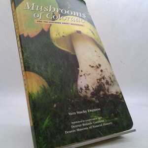 May include: A book titled "Mushrooms of Colorado" with a photograph of mushrooms on the cover. The cover features a large mushroom with a yellow cap and white stem, set against a backdrop of green grass and foliage. The author's name is Vera Stucky Evenson.