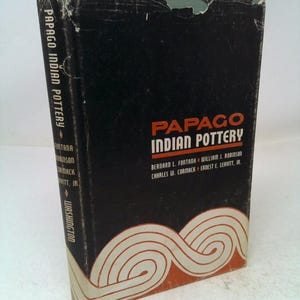 May include: A hardcover book titled Papago Indian Pottery with a black dust jacket. The cover features orange text and a white graphic of swirling lines at the bottom. The spine displays the title and authors, showing signs of wear at the top edge.