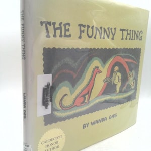 May include: A hardcover book titled The Funny Thing by Wanda Gág with a pale yellow cover. The front features a dark rectangular illustration of a red creature and a person holding a plate. A Caldecott Honor Author seal is printed on the cover.