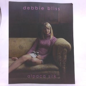 May include: A promotional image for Debbie Bliss alpaca silk. The image features a woman wearing a light purple cable-knit sweater, seated on a vintage-style sofa. The text "debbie bliss" and "alpaca silk" are displayed at the top and bottom of the image.