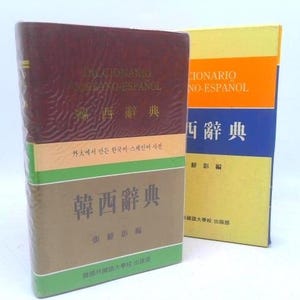 May include: Two dictionaries, one burgundy and the other yellow, stand side by side. The burgundy dictionary has text in multiple languages, including Korean and Spanish. The yellow dictionary has orange and blue sections with Spanish text.