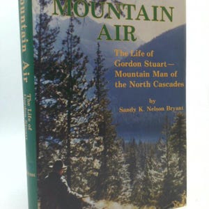 May include: A vintage book titled "Mountain Air" with a forest scene on the cover. The title is in large green letters, with the subtitle "The Life of Gordon Stuart - Mountain Man of the North Cascades" below. The author's name is Sandy K. Nelson Bryant.