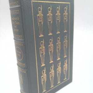 May include: A hardcover book with a dark green cover and gold accents. The cover features a gold illustration of soldiers. The spine has gold lettering that reads "Andersen's Fairy Tales and Stories". A gold bookmark is visible.