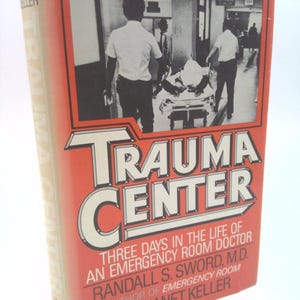 May include: A book cover with the title "Trauma Center" in large, bold, white letters on a red background. The subtitle is "Three Days in the Life of an Emergency Room Doctor" by Randall S. Sword, M.D. and Janet Keller. The cover features a black and white photo of medical personnel pushing a stretcher with a patient.