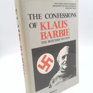 May include: A hardcover book titled "The Confessions of Klaus Barbie, The Butcher of Lyon." The cover features the title in black and red, a portrait, and a red swastika symbol on a black background. The author's name is Robert Wilson.