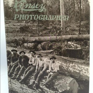 May include: A black and white photograph of five men sitting on a large fallen log in a forest. The men are wearing work clothes and hats. The text "Kinsey Photographer" is printed at the top of the image.