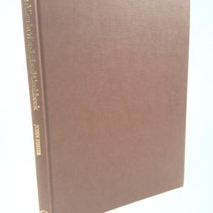 May include: A hardcover cookbook titled "Alice in Wonderland Cookbook" by John Fisher. The book has a brown cover with gold lettering on the spine. The cover has a textured appearance.