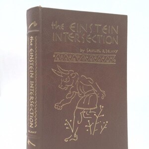 May include: A hardcover book titled "The Einstein Intersection" by Samuel R. Delany. The cover is brown with gold lettering and a gold illustration of a figure. The spine also features the title and author's name.