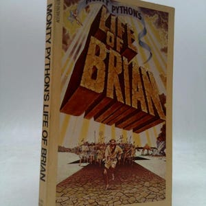 May include: A paperback book titled Monty Python's Life of Brian with a tan cover. The front features a large, cracked, three-dimensional title floating above a desert scene with a running figure and a crowd of people under a bright sun.