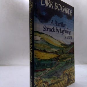 May include: A hardcover book titled A Postillion Struck by Lightning, A Memoir by Dirk Bogarde. The dust jacket features a watercolor-style landscape painting with rolling green hills, yellow fields, and a dark blue sky. The spine is black.
