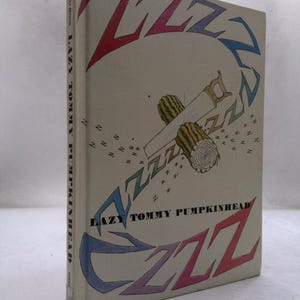 May include: A hardcover book titled Lazy Tommy Pumpkinhead by Pène du Bois. The off-white cover features a central illustration of a hand saw cutting through a log, surrounded by large, colorful, stylized ZZZ letters and smaller scattered Zs.