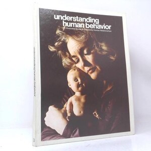 May include: A hardcover book titled Understanding Human Behavior: An Illustrated Guide to Successful Human Relationships. The cover features a dark-toned photograph of a person holding a small doll, with white text printed at the top of the cover.