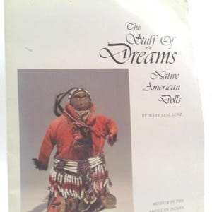 May include: A book cover featuring a Native American doll and the title "The Stuff of Dreams: Native American Dolls." The doll is wearing a red garment and beaded accessories. The book is by Mary Jane Lenz.