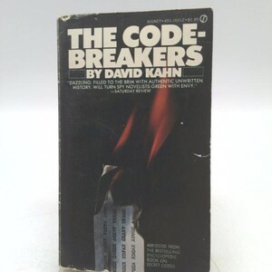 May include: A black and white paperback book titled "The Code-Breakers" by David Kahn. The book cover features a red and orange abstract design. The text "DAZZLING. FILLED TO THE BRIM WITH AUTHENTIC UNWRITTEN HISTORY, WILL TURN SPY NOVELISTS GREEN WITH ENVY." is printed on the cover. The text "SATURDAY REVIEW" is printed below the quote.