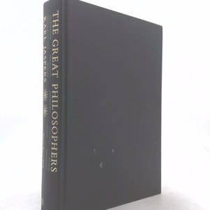 May include: A black hardcover book titled The Great Philosophers by Karl Jaspers. The spine features gold lettering and two small star-like symbols. The book is positioned at an angle, showing the front cover and the spine clearly against a white background.