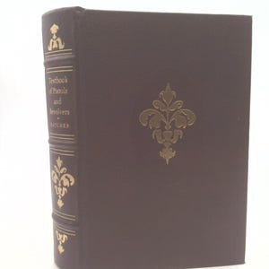 May include: A brown hardcover book titled "Textbook of Pistols and Revolvers" by Hatcher. The book has gold decorative accents on the spine and front cover. The book is standing upright.