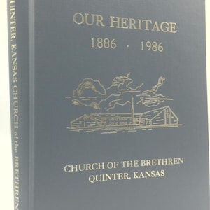 May include: A blue book titled "OUR HERITAGE" with gold lettering and a gold illustration of a building. The book's spine reads "QUINTER, KANSAS CHURCH of the BRETHREN". The years 1886 and 1986 are also displayed.