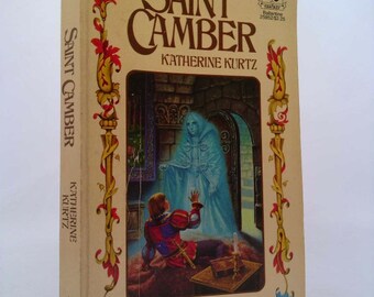 May include: A book cover with a yellow background and a title that reads "Saint Camber" by Katherine Kurtz. The cover features an illustration of a man in a red and blue tunic kneeling before a ghostly figure in a white gown. The text "Volume II in the Legends of Camber of Culdi" is printed at the bottom of the cover.