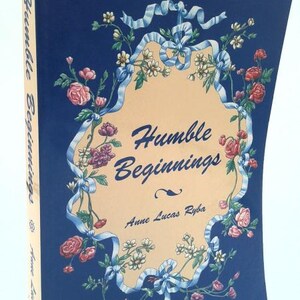 May include: A hardcover book titled "Humble Beginnings" by Anne Lucas Ryba. The cover is navy blue with a floral design in pink, white, and blue. The title is in cursive, and the spine also has the title.