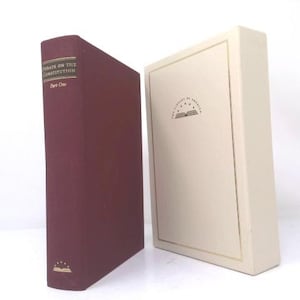 May include: A maroon hardcover book titled Debate on the Constitution Part One with gold lettering on the spine, positioned next to a cream-colored slipcase featuring the Library of America logo and a gold-lined rectangular border on the front cover.
