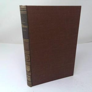 May include: A brown hardcover book with a dark spine featuring gold lettering that reads Plotinus, the number 17, a small emblem, and the text Great Books. The book has a textured fabric cover and is standing upright on its bottom edge.