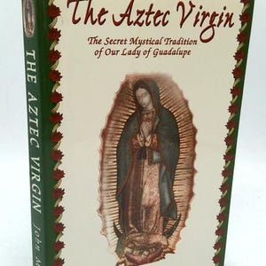 May include: A hardcover book titled "The Aztec Virgin" with a cream-colored cover featuring an illustration of a religious figure. The book's spine is green with the title in white. The cover also includes the text "The Secret Mystical Tradition of Our Lady of Guadalupe."