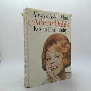 May include: A vintage book titled "Always Ask a Man Arlene Dahl's Key to Femininity." The cover features a portrait of a woman with red hair and the title in red and black text. The book is off-white with some wear.