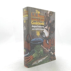 May include: The L.L. Bean Game & Fish Cookbook by Angus Cameron and Judith Jones. The cover features a rustic illustration of a cooking pot, vegetables, and fish. The book is a hardcover with a wooden-like texture.
